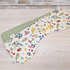 May include: A curved burp cloth featuring a cream-coloured fabric with a botanical print of red, yellow, and blue flowers, green leaves, and small mushrooms. The reverse side is a solid sage green fabric with two small white brand tags attached.
