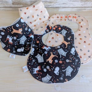 May include: Three cotton baby bibs with different patterns. One bib is black with a white cat print, another is beige with brown paw prints, and the third is beige with brown paw prints and a white cat print.