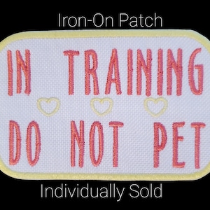 May include: Rectangular iron-on patch with a white background and a yellow border. The patch features the text "IN TRAINING DO NOT PET" in coral-colored, embroidered letters. Three small yellow hearts are embroidered between the lines of text.
