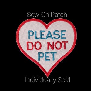 May include: A heart-shaped sew-on patch with a white center, red border, and the words "PLEASE DO NOT PET" in blue and red. The patch is against a black background.