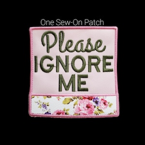 May include: A square, pink sew-on patch with the words "Please IGNORE ME" embroidered in green. The bottom features a floral pattern with pink roses and purple flowers. The patch is outlined in pink.
