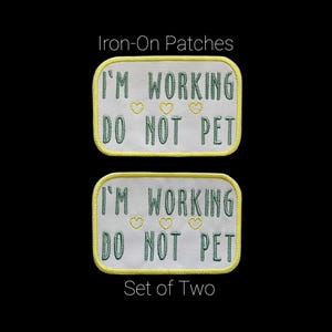 May include: Two rectangular iron-on patches with rounded corners. Each patch has the text "I'M WORKING DO NOT PET" embroidered in green thread. Three small yellow hearts are embroidered above the text. The patches are outlined in yellow.