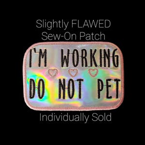 May include: A rectangular sew-on patch with a holographic background and a pink border. The patch reads "I'M WORKING DO NOT PET" in black text, with three embroidered hearts. The text "Slightly FLAWED" and "Individually Sold" are also present.