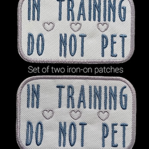 May include: Two rectangular iron-on patches with the text "IN TRAINING DO NOT PET" in blue, with three small heart shapes. The patches have a light gray border and are on a black background.