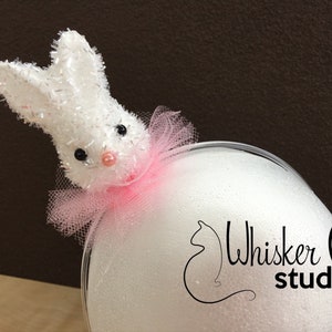 May include: A white bunny decoration with a pink tulle collar and black bead eyes. The bunny is made of a fuzzy material and is perched on a white, rounded surface. The image includes the text "Whisker Pinch studio".