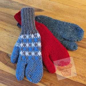 Puede incluir: Tres mitones de punto en tonos azul, rojo y gris. Un mitón azul presenta un patrón geométrico en blanco y gris. El mitón rojo es liso y el mitón gris es liso. Los mitones están colocados sobre una superficie de madera.