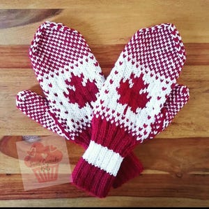 May include: A pair of knitted mittens featuring a red and white maple leaf pattern. The mittens have a ribbed cuff with alternating red and white stripes. A small square label with the text Sweet Cupcake Knits is visible.