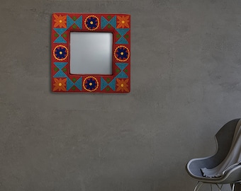 Op de afbeelding: Een vierkante spiegel met een kleurrijk, handgeschilderd frame. Het frame heeft een geometrisch patroon met rode, blauwe, gele en oranje accenten. De spiegel hangt aan een grijze muur in een kamer met een stoel en een raam.
