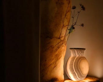 May include: A glowing, decorative vase with a unique, layered pattern. The vase is illuminated from within, casting a warm light. It sits on a dark, textured surface, with a dried plant stem in the background.
