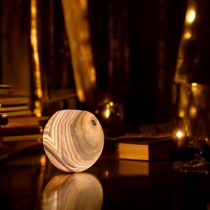 Peut inclure: Une lampe ronde en bois avec un motif rayé brun clair, beige et gris. La lampe est posée sur une surface sombre.