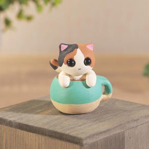 Puede incluir: Figura de cerámica artesanal de un gato calicó asomando de una taza de té turquesa. El gato tiene pelaje negro, marrón y blanco con grandes ojos negros. La taza es turquesa con base y asa beige. La figura está sobre un bloque de madera.