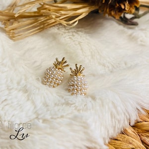 Könnte beinhalten: Ein Paar goldene Ananas-Ohrringe mit weißen Perlenakzenten. Die Ohrringe liegen auf einer weißen, flauschigen Oberfläche.