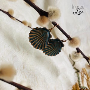 Könnte beinhalten: Türkise und kupferfarbene Muschel-Ohrringe mit filigranem Design. Die Ohrringe hängen an einem Zweig mit flauschigen weißen Knospen.