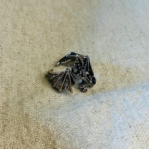 Könnte beinhalten: Ein silberfarbener Drachenring mit detaillierten Flügeln und Körper. Der Ring ist offen, um die Größe anzupassen. Die Flügel des Drachen sind ausgebreitet, und das Design ist aufwendig, mit einem Fantasy-Thema.