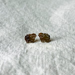 Könnte beinhalten: Ein Paar goldfarbene Ohrstecker in Form von Schafen. Jeder Ohrring hat eine detaillierte Umrisszeichnung eines Schafes mit flauschigem Körper und kleinem Gesicht. Auf einem weißen, strukturierten Hintergrund.