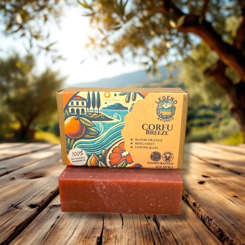 May include: A bar of handmade soap with a red-brown color and a rough texture. The soap is packaged in a cardboard box with a colorful illustration of a Mediterranean island scene and the text "Legend Naturals Corfu Breeze Blood Orange Bergamot Lemongrass Handcrafted 4 oz (113g)".