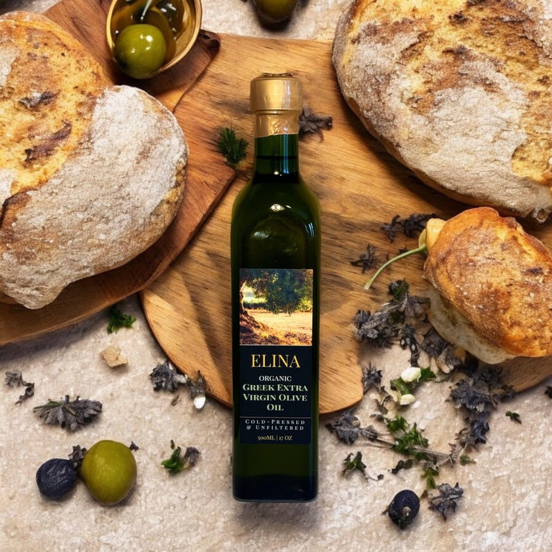 May include: A green glass bottle of "ELINA" Organic Greek Extra Virgin Olive Oil, 500ml (17 fl oz), with a gold cap, sits on a wooden cutting board. The image includes two loaves of bread, olives, and sprigs of herbs.