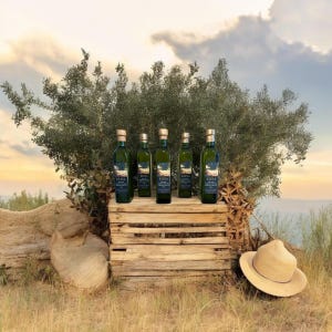May include: Five green glass bottles of olive oil with gold-colored caps are displayed on a rustic wooden crate. The bottles are labeled with dark blue labels. A straw hat rests on the ground in front of the crate, with an olive tree in the background.
