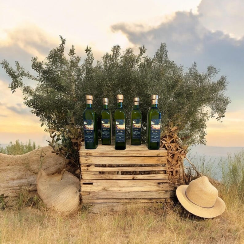 May include: Five green glass bottles of olive oil with gold-colored caps are displayed on a rustic wooden crate. The bottles are labeled with dark blue labels. A straw hat rests on the ground in front of the crate, with an olive tree in the background.