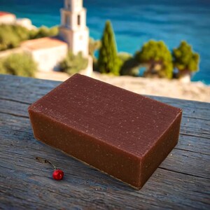 May include: A rectangular bar of brown soap with a grainy texture, sitting on a wooden surface with a red cherry in the foreground.