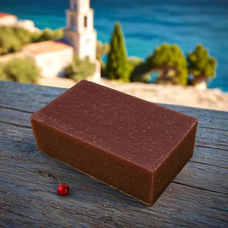 May include: A rectangular bar of brown soap with a grainy texture, sitting on a wooden surface with a red cherry in the foreground.