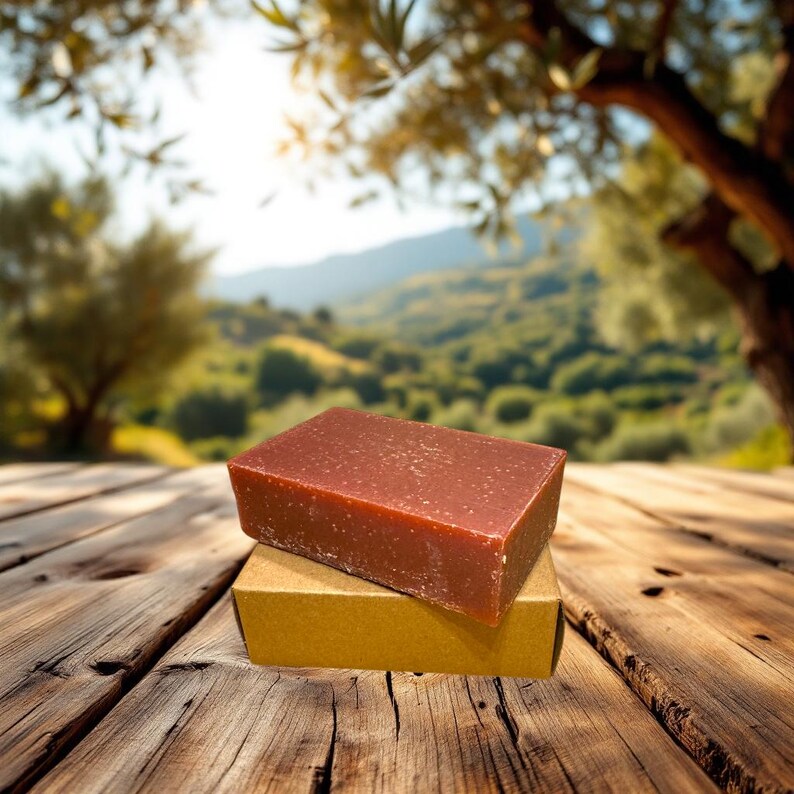 May include: A brown bar of soap with a rough texture, sitting on a gold cardboard box. The soap is on a wooden surface with a blurred background of green trees and a blue sky.
