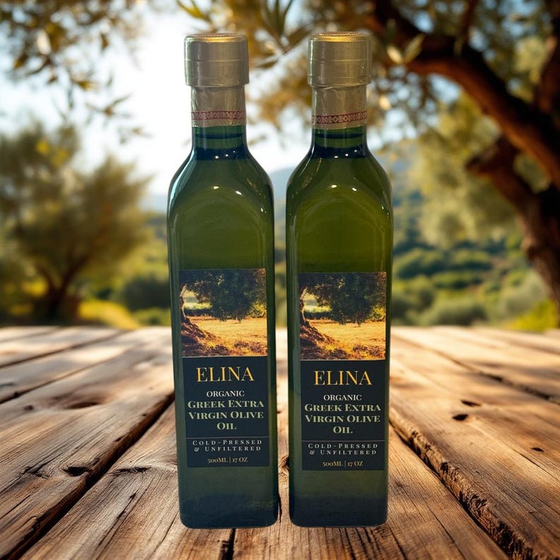 May include: Two green glass bottles of Elina Organic Greek Extra Virgin Olive Oil. Each bottle has a gold cap and a label with the brand name and product details. The bottles are on a wooden surface with a blurred outdoor background.
