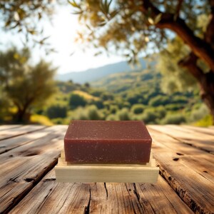 May include: A bar of dark brown soap on a wooden soap dish. The soap is sitting on a wooden table with a blurred background of green hills and trees.