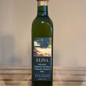 May include: A green glass bottle of Elina Organic Greek Extra Virgin Olive Oil. The label features a landscape image and text. The bottle is 17 oz and has a gold foil seal on the top.