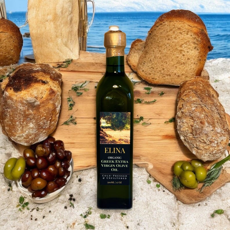 May include: A green glass bottle of Elina Organic Greek Extra Virgin Olive Oil, with a gold cap, sits on a wooden board. The label features a landscape scene. Surrounded by fresh bread, olives, and herbs, the scene evokes a rustic, Mediterranean feel.