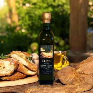 May include: A green glass bottle of Elina Organic Greek Extra Virgin Olive Oil, with a gold cap, sits on a wooden surface next to bread and a cup of olive oil. The label features a tree illustration and the text "COLD-PRESSED & UNFILTERED".