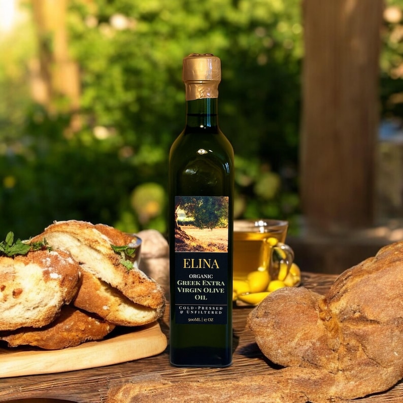 May include: A green glass bottle of Elina Organic Greek Extra Virgin Olive Oil, with a gold cap, sits on a wooden surface next to bread and a cup of olive oil. The label features a tree illustration and the text "COLD-PRESSED & UNFILTERED".