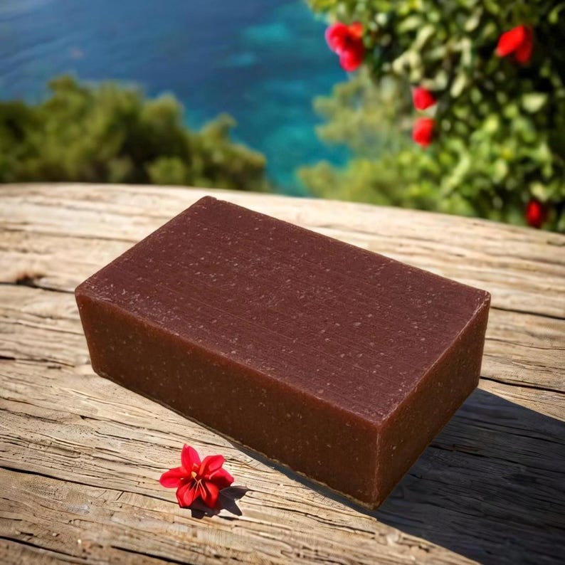 May include: A rectangular bar of dark brown soap with a grainy texture, resting on a wooden surface. A small red flower is in the foreground.