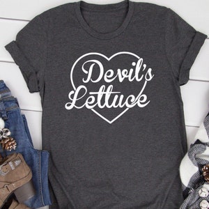 Puede incluir: Una camiseta gris oscura con un diseño de corazón blanco que dice "Devil's Lettuce" en una fuente blanca cursiva.
