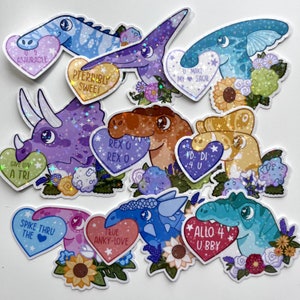 May include: A set of ten holographic dinosaur stickers with glitter and floral accents. Each sticker features a different dinosaur with a heart-shaped message. The messages include "U Make My Saur", "Pterribly Sweet", "Rex O Rex O", "#D Di 4 U", "Give Luv A Tri", "Spike Thru The", "True Anky-Love", and "Allo 4 U Bby".