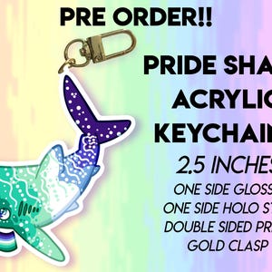 Puede incluir: Un llavero acrílico Pride Shark con cierre dorado. El tiburón presenta un diseño colorido con una bandera y el texto "PRIDE SHARK ACRYLIC KEYCHAINS". El llavero mide 6,35 cm y tiene un acabado brillante y holográfico.