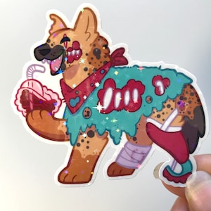 May include: A cartoon sticker of a hyena with brown and black spots, wearing a red bandana and a green and blue tattered cloak. The hyena is holding a red and brown cup with a straw and has a bandage on its leg. The sticker has a glittery finish.
