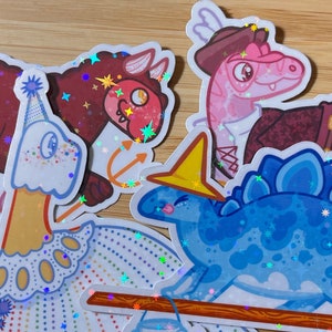 May include: Four colorful holographic stickers featuring cartoon dinosaurs. The stickers are on a wooden surface. The dinosaurs are a red and brown dinosaur with an anchor, a blue dinosaur with a yellow crown, a pink dinosaur wearing a pirate hat and a white dinosaur wearing a party hat.