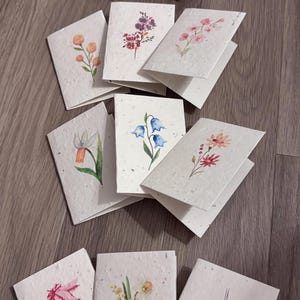 May include: A collection of folded greeting cards made from off-white, textured paper with visible flecks. Each card features a unique watercolour illustration of various flowers, including bluebells, daffodils, and bird of paradise blossoms.