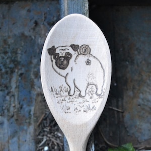 May include: A wooden spoon with a black and white image of a pug dog burned into the surface. The pug is facing the left side of the spoon and has a flower on its back.
