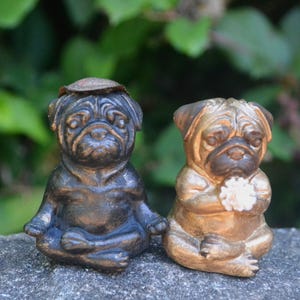 May include: Two pug figurines in a seated meditation pose. The left pug is dark bronze with a small flat cap on its head. The right pug is a lighter metallic gold color and holds a small white flower-like object between its front paws.