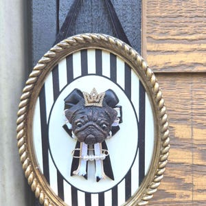 May include: An oval wall hanging featuring a black and white striped background inside a gold rope-textured frame. A sculpted black pug head wearing a small gold crown sits atop a black and white ribbon rosette with a safety pin and white beads.