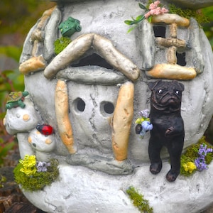 May include: A whimsical, gray stone fairy house with a pug figurine holding flowers. The house features mushroom accents, a ladybug, and a turtle. The scene is set outdoors with greenery in the background.