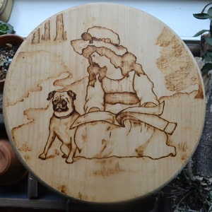 May include: Round wooden plaque with a pyrography design. The artwork depicts a woman in a hat and dress, seated with a pug dog. The design is burned into the wood, creating a light brown image on a lighter wood background.