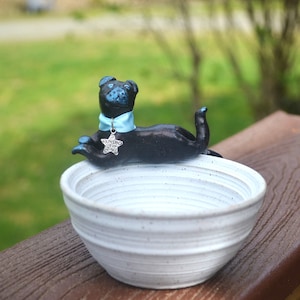 May include: A ceramic dog figurine in black and blue, wearing a light blue collar and a star-shaped tag that reads "Just for you." The dog rests atop a white, textured ceramic bowl, perfect for a pet's food or water.