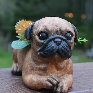 May include: A brown pug figurine with a black muzzle and ears, adorned with an orange and green floral accent. The pug is in a seated position with its paws crossed, creating a charming and decorative piece.