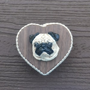 May include: A heart-shaped decorative item featuring a pug's face. The pug's face is cream and black, set against a dark wood background. The heart is outlined with a white rope-like trim.