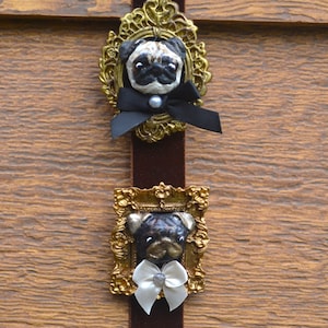 May include: Two pug dog head sculptures mounted in ornate gold-colored frames attached to a vertical brown velvet ribbon. The top pug features a black ribbon bow with a pearl accent, while the bottom pug features a white ribbon bow with a bead.