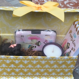 May include: A yellow gift box with a bee pattern, containing a phone case, notebook, candle, and a small black item with a pink bow. The notebook reads "Stay Pawsitive". A yellow bow adorns the box.