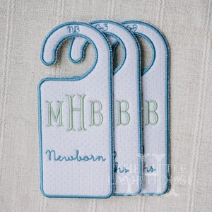May include: Three white fabric door hangers with blue trim and polka dots. The first hanger has the initials "MHB" in green and the word "Newborn" in blue script. The other two hangers have the letter "B" in green and the numbers "0-3" and "3-6" in blue.